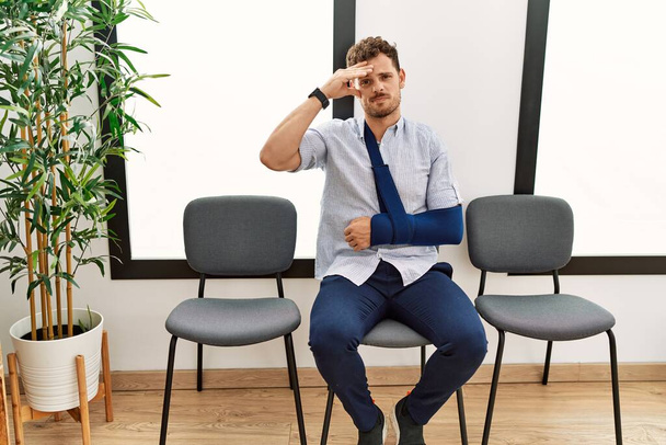 Schöner junger Mann, der mit Armverletzung im Wartezimmer des Arztes sitzt, besorgt und gestresst über ein Problem mit der Hand auf der Stirn, nervös und krisenbesorgt  - Foto, Bild