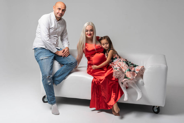 Hermosa madre embarazada en un vestido rojo y padre con una chica linda y emoticono en un sofá blanco aislado sobre un fondo blanco. Familia, concepto de nacimiento del bebé - Foto, Imagen