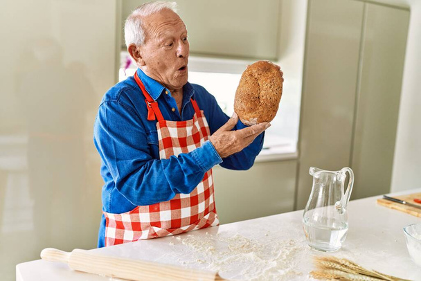 Senior man smiling confident holding wholemeal bread at kitchen - Fotografie, Obrázek