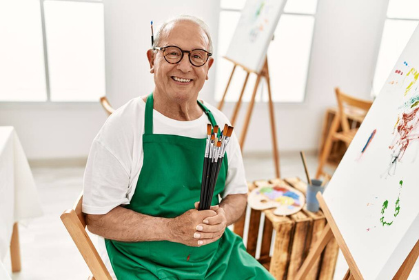 Älterer grauhaariger Künstler lächelt glücklich und hält Pinsel im Kunstatelier. - Foto, Bild