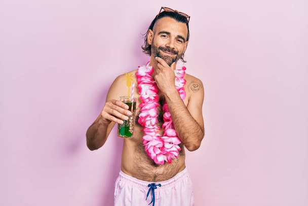 Young hispanic man wearing swimsuit and hawaiian lei drinking tropical cocktail looking confident at the camera smiling with crossed arms and hand raised on chin. thinking positive.  - Photo, Image