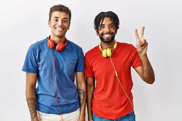 Młodzi latynoscy bracia stojący nad odosobnionym tłem, noszący słuchawki pokazujące i wskazujące palcami numer dwa, uśmiechający się z pewnością siebie i szczęściem..  - Zdjęcie, obraz