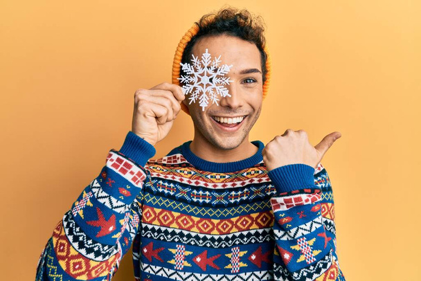 Νεαρός όμορφος άντρας κρατώντας νιφάδες χιονιού φορώντας χειμωνιάτικο πουλόβερ δείχνοντας τον αντίχειρα στο πλάι χαμογελώντας χαρούμενος με ανοιχτό στόμα  - Φωτογραφία, εικόνα