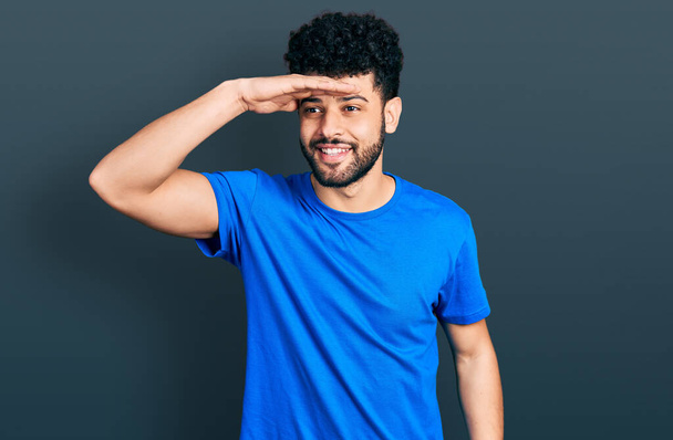 Hombre árabe joven con barba vistiendo camiseta azul casual muy feliz y sonriente mirando lejos con la mano sobre la cabeza. concepto de búsqueda.  - Foto, imagen