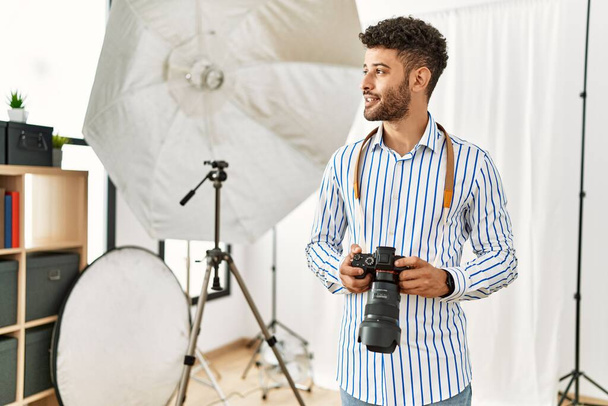 Νεαρός Άραβας φωτογράφος χαμογελά χαρούμενος χρησιμοποιώντας αντανακλαστική κάμερα στο φωτογραφικό στούντιο. - Φωτογραφία, εικόνα