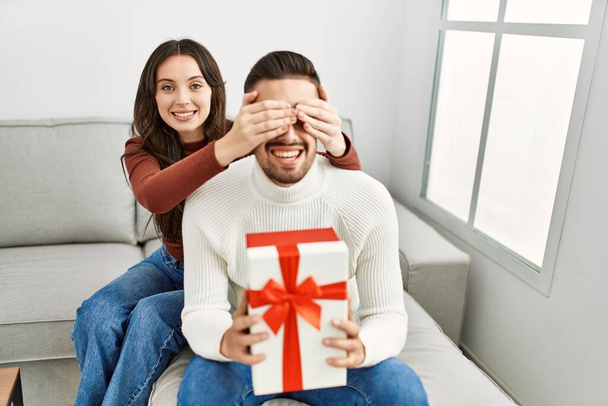 Νεαρό ζευγάρι ισπανόφωνων χαμογελά ευτυχής έκπληξη με δώρο γενεθλίων στο σπίτι. - Φωτογραφία, εικόνα