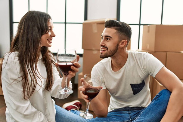 Joven pareja hispana sonriendo feliz brindis con vino tinto en nuevo hogar. - Foto, imagen