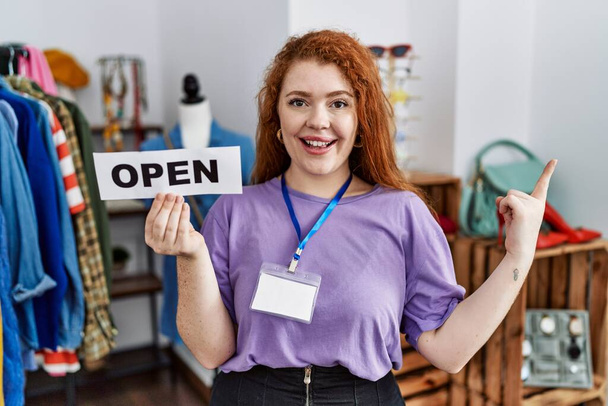 Young redhead woman holding banner with open text at retail shop smiling happy pointing with hand and finger to the side  - Φωτογραφία, εικόνα