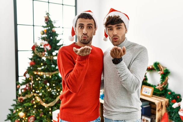 Young gay couple standing by christmas tree wearing hat looking at the camera blowing a kiss with hand on air being lovely and sexy. love expression.  - Photo, Image