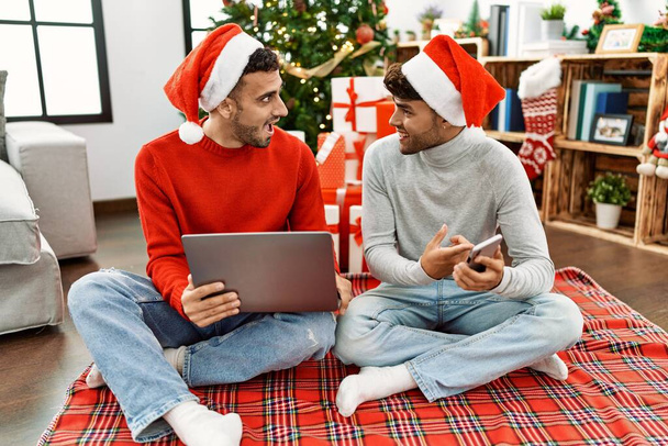 Dva hispánští muži pár pomocí notebooku a smartphone sedí u vánočního stromku doma - Fotografie, Obrázek