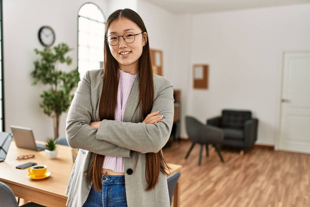 Joven mujer de negocios china sonriendo feliz de pie con los brazos cruzados gesto en la oficina. - Foto, Imagen