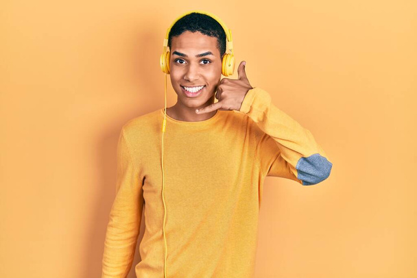Young african american guy listening to music using headphones smiling doing phone gesture with hand and fingers like talking on the telephone. communicating concepts.  - 写真・画像