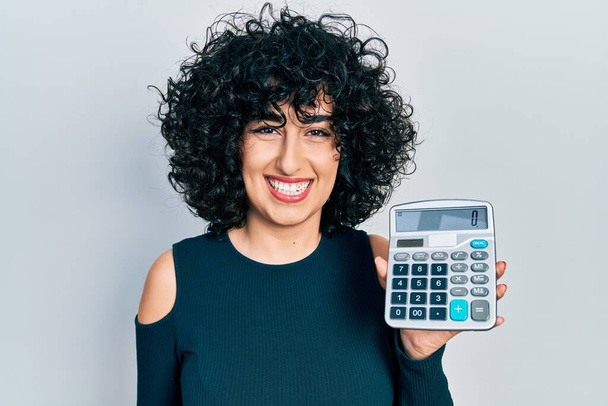Young middle east woman showing calculator device looking positive and happy standing and smiling with a confident smile showing teeth  - Foto, Bild