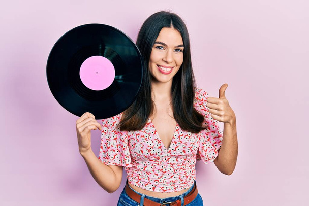 Mladá brunetka žena drží vinyl disk s úsměvem šťastný a pozitivní, palec nahoru dělá vynikající a schvalovací znamení  - Fotografie, Obrázek