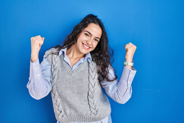 Young brunette woman standing over blue background celebrating surprised and amazed for success with arms raised and eyes closed. winner concept.  - 写真・画像