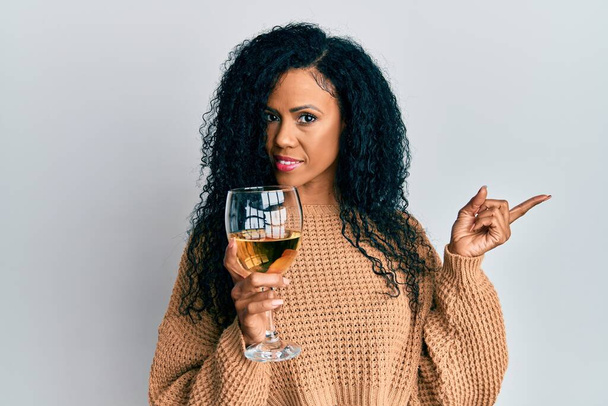 Middle age african american woman drinking a glass of white wine smiling happy pointing with hand and finger to the side  - Foto, immagini