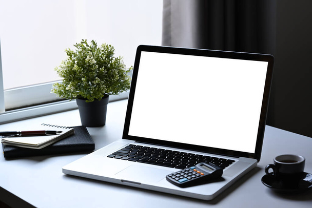 Φορητός υπολογιστής, φλιτζάνι καφέ, σημειωματάριο και αριθμομηχανή σε λευκό τραπέζι. Κενή οθόνη για μοντάζ γραφικών προβολών. - Φωτογραφία, εικόνα