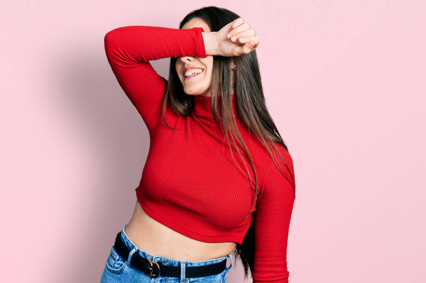 Young brunette teenager wearing red turtleneck sweater covering eyes with arm smiling cheerful and funny. blind concept.  - Fotografie, Obrázek