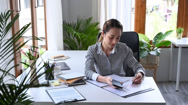 Вдумчивая молодая женщина-предприниматель держит смартфон и проверяет финансовые отчеты. - Фото, изображение