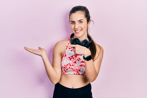 Young brunette teenager wearing gym clothes and using headphones amazed and smiling to the camera while presenting with hand and pointing with finger.  - 写真・画像