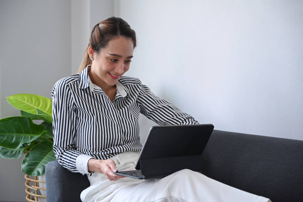 Szczęśliwa młoda kobieta siedzi na kanapie w domu i ogląda filmy, surfuje po Internecie za pomocą tabletu cyfrowego. - Zdjęcie, obraz