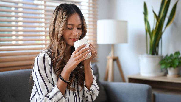 Relaxed young woman sitting on sofa and drinking hot coffee in the morning. - Foto, Bild