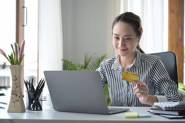 Smiling millennial woman holding credit card and using internet banking service on laptop computer. - Photo, Image