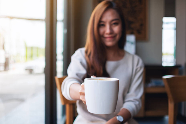 Closeup image of a beautiful young asian woman holding and giving a cup of hot coffee in cafe - Foto, imagen