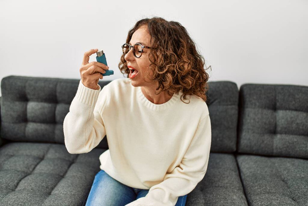 Mujer hispana madura usando inhalador de asma sentada en el sofá en casa - Foto, imagen