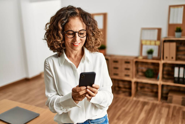 Middelbare leeftijd Spaanse vrouw glimlachen vol vertrouwen met behulp van smartphone op kantoor - Foto, afbeelding