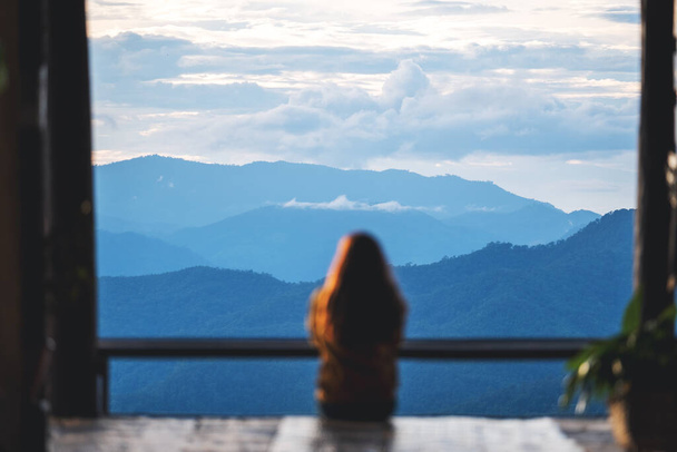 Blurred rear view image of a female traveler sitting and looking at a beautiful mountain and nature view before sunset - Foto, Imagen