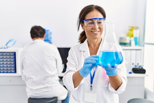 Middelbare leeftijd man en vrouw partners dragen wetenschapper uniform houden reageerbuis in laboratorium - Foto, afbeelding