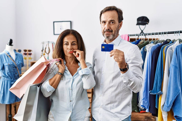 Hispanisches Paar mittleren Alters mit Einkaufstaschen und Kreditkartenmund und -lippen als Reißverschluss mit Fingern. Geheimniskrämerei und Schweigen, Tabuthema  - Foto, Bild