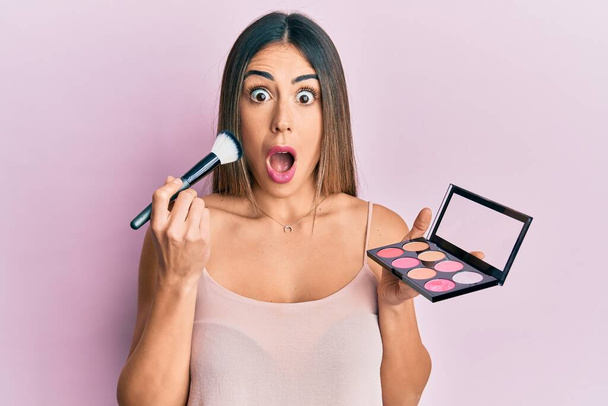 Young hispanic woman holding makeup brush and blush afraid and shocked with surprise and amazed expression, fear and excited face.  - Photo, Image