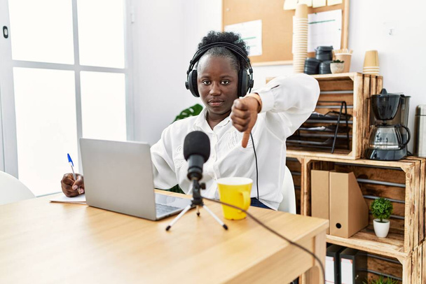 Νεαρή Αφρικανή που δουλεύει σε ραδιοφωνικό στούντιο με θυμωμένο πρόσωπο, αρνητικό σημάδι που δείχνει απέχθεια με τους αντίχειρες κάτω, απορριπτική ιδέα  - Φωτογραφία, εικόνα