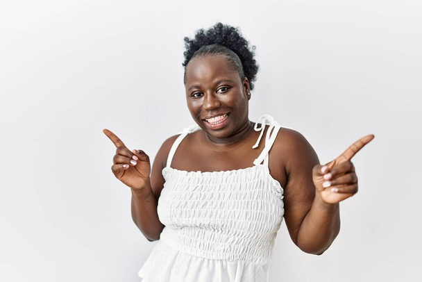 Mladá africká žena, stojící nad bílým izolovaným pozadím a usmívající se sebevědomě ukazujíc prstem do různých směrů. kopírovat místo pro inzerci  - Fotografie, Obrázek