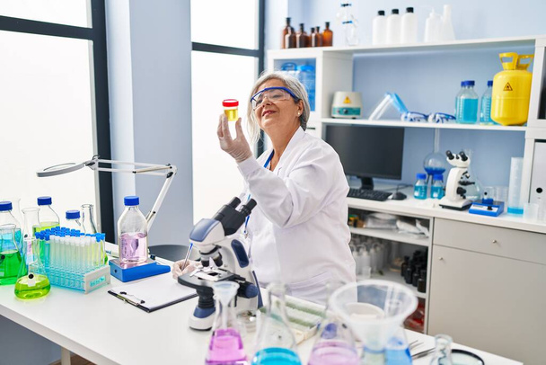 Женщина средних лет в униформе ученого держит пробирку для анализа мочи в лаборатории - Фото, изображение