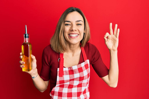 Jeune femme blonde caucasienne portant tablier de cuisinier tenant bouteille d'huile d'olive souriant et riant fort à haute voix parce que drôle blague folle.  - Photo, image