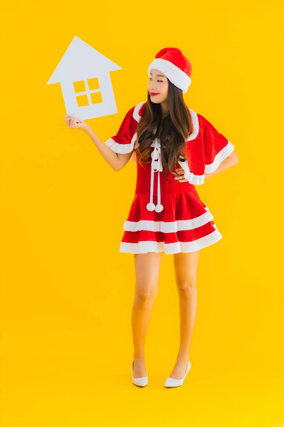 Portret mooie jonge aziatische vrouw dragen kerstmis kleding en hoed show huis huis teken met veel geld en geld op gele geïsoleerde achtergrond - Foto, afbeelding