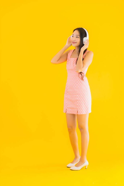 Retrato hermosa joven asiática mujer escuchar música con el teléfono móvil inteligente y auriculares en amarillo aislado fondo
 - Foto, Imagen