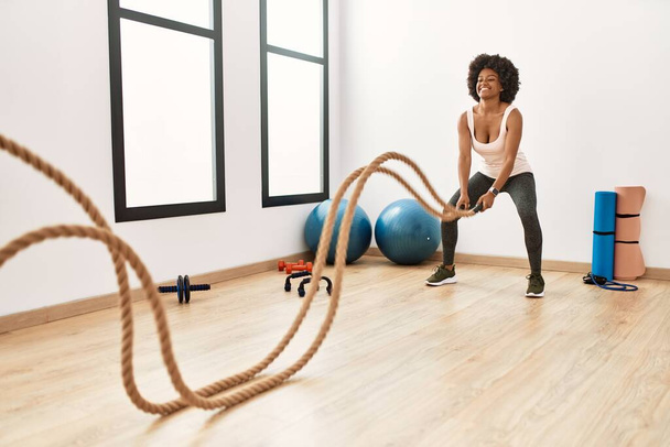Молодая африканская американка улыбается уверенной в себе тренировкой с боевой веревкой в спорткомплексе - Фото, изображение