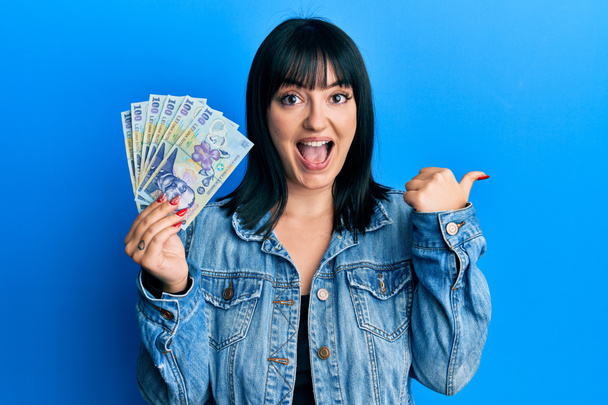 Joven mujer hispana sosteniendo 100 billetes de leu rumanos apuntando con el pulgar hacia un lado sonriendo feliz con la boca abierta  - Foto, Imagen