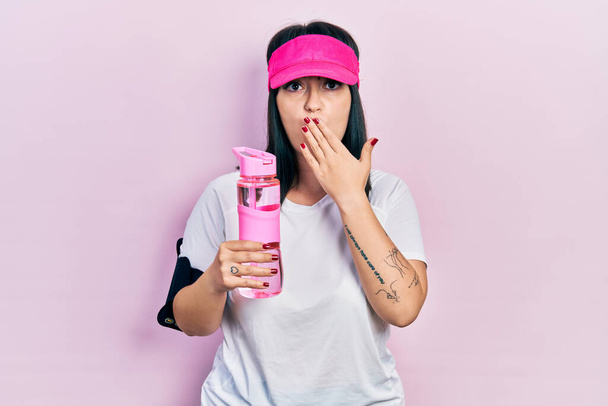 Junge hispanische Frau in Sportkleidung, die eine Flasche Wasser trinkt und den Mund mit der Hand bedeckt, schockiert und aus Angst vor Fehlern. Überraschte Miene  - Foto, Bild