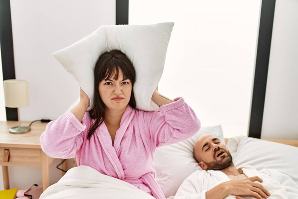 Hispanic woman angry with pillow on his ears while man snoring on the bed. - Photo, Image