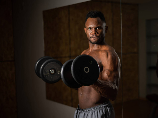 Muscular dark-skinned man doing an exercise with dumbbells.  - Zdjęcie, obraz