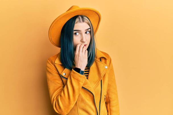 Młoda nowoczesna dziewczyna w żółtym kapeluszu i skórzanej kurtce wyglądająca na zestresowaną i zdenerwowaną z rękami obgryzającymi paznokcie. niepokój.  - Zdjęcie, obraz