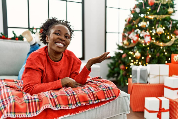 Młoda Afroamerykanka leżąca na kanapie przy choince uśmiecha się radośnie prezentując i wskazując dłonią na aparat fotograficzny.  - Zdjęcie, obraz