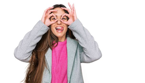Una joven hispana vestida con ropa de negocios haciendo un buen gesto como binoculares sacando la lengua, ojos mirando a través de los dedos. expresión loca.  - Foto, imagen
