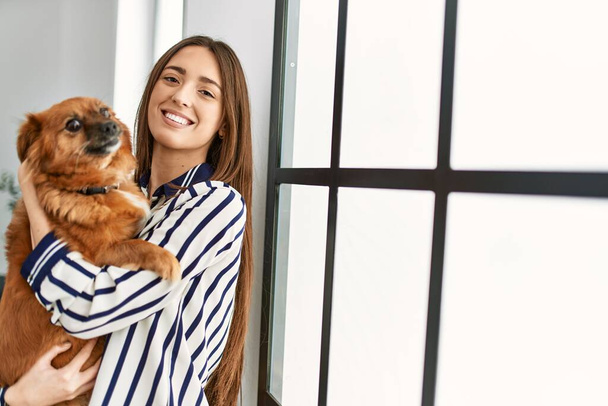 Молода іспаномовна жінка посміхається впевнено обіймає собаку, що стоїть вдома
 - Фото, зображення