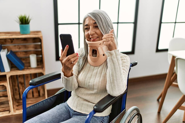 Grauhaarige Frau mittleren Alters, die zu Hause im Rollstuhl sitzt und Telekonsultation hat - Foto, Bild